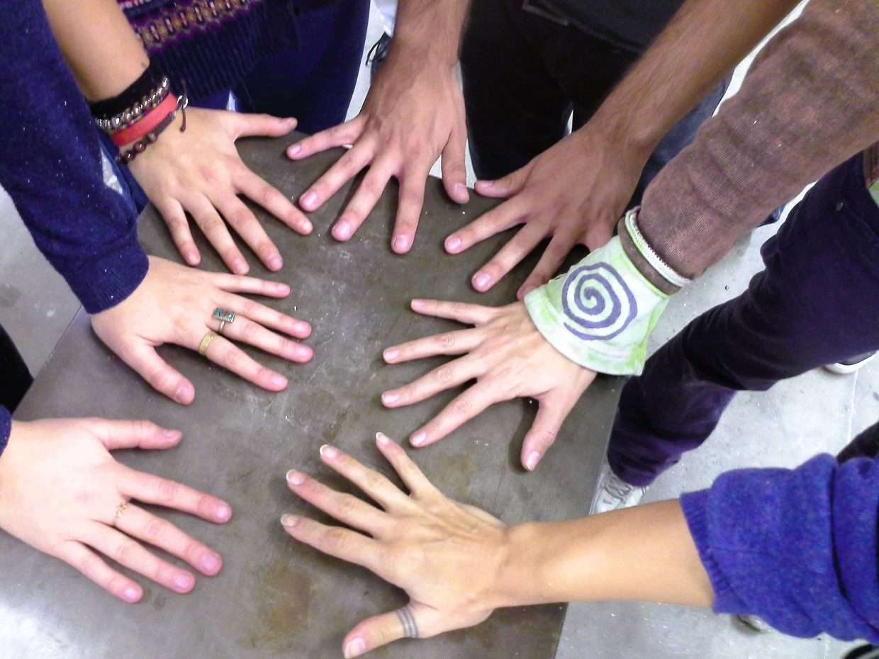 Nella foto a colori di formato rettangolare si vedono sette mani di ragazzi appoggiate su un piano di legno grigio.