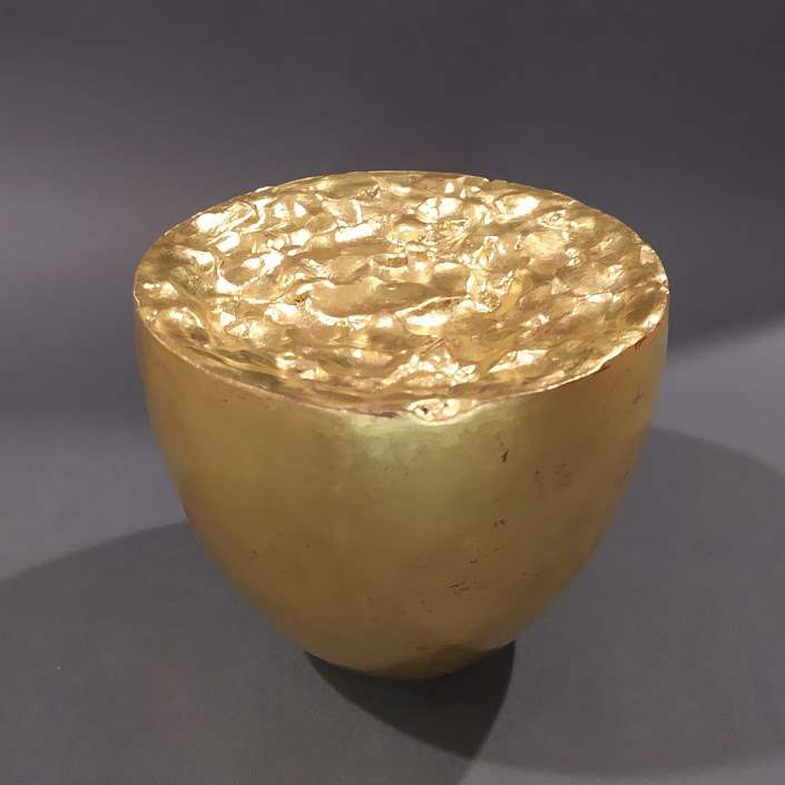 Nell'immagine di formato quadrato si vede al centro, su di uno sfondo grigio scuro, una scultura dorata a forma di tazza piena fino all'orlo di una specie di liquido plastico in movimento.