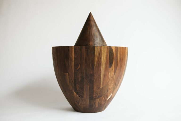 Nell'immagine si vede una scultura in legno a forma di vaso con una forma a punta sulla sommità
